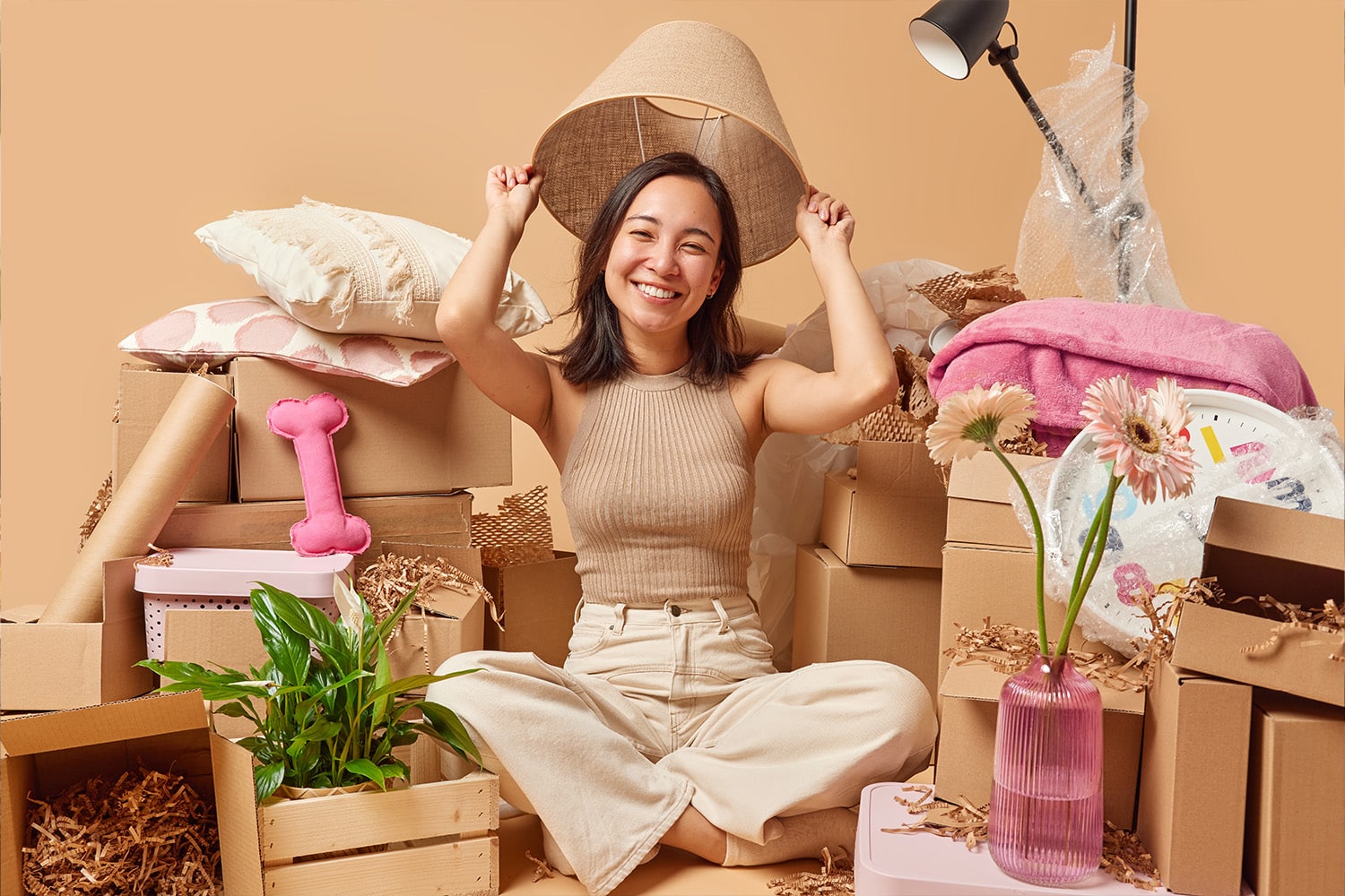 Positive young Asian woman poses under lampshade sits crossed legs on floor relocates to new place of living surrounded by cardboard boxes isolated over brown background. Moving Day concept